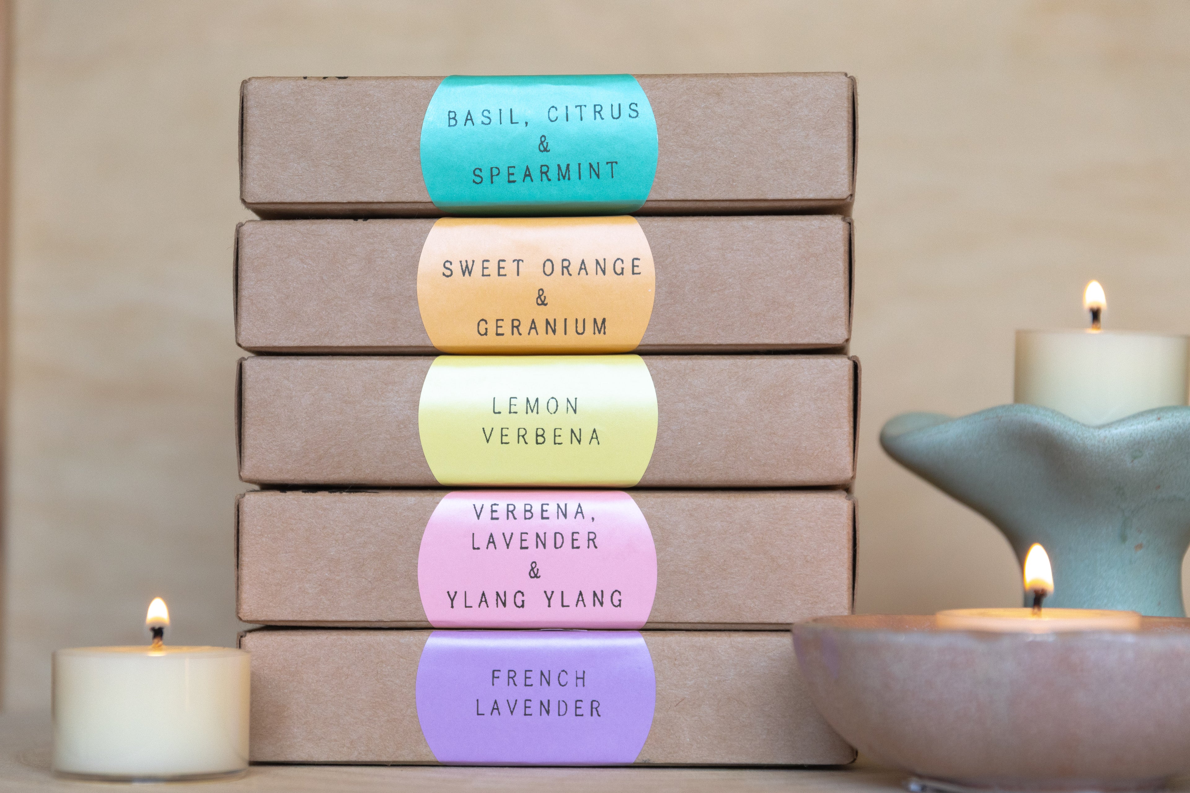 Stack of tealight boxes with colorful labels noting each scent and displayed lit tealight candles on a neutral background