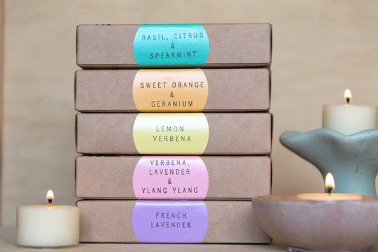 Stack of tealight boxes with colorful labels noting each scent and displayed lit tealight candles on a neutral background