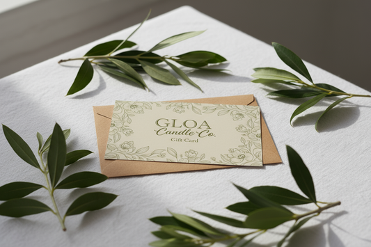Please can you create me an image of a gift card, it should have the words 'GLOA Candle Co' in bigger text, with smaller text underneath which reads 'Gift Card'. The card should be colour #F5F2E9, with #8e8b63 green floral print on it, it should be displayed on a table with white cloth, a brown envelope and green leaves around